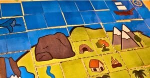 A colorful board game map with illustrated tiles showing land and water forms—water, rocks, mountains, a house, a campfire, trees, and a boat—perfect for exploring island activities. Montessori Nature Printables