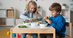 Two children sit at a small table, each peeling green peas and placing them into white bowls. Immersed in the rhythm of work, they demonstrate a child's cycle of concentration as they focus intently on their task. The background shows shelves with toys and decorations in a brightly lit room. Montessori Nature Printables