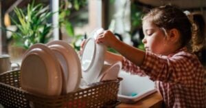 A young, capable girl places clean plates into a drying rack beside a kitchen sink, with plants visible in the background—showcasing how real work can be done gracefully by children. Montessori Nature Printables