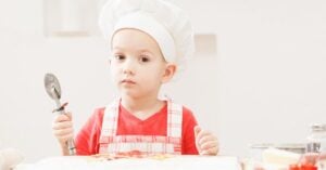A young Montessori toddler in a chef's hat, practicing practical life skills in the kitchen as he holds a spoon.