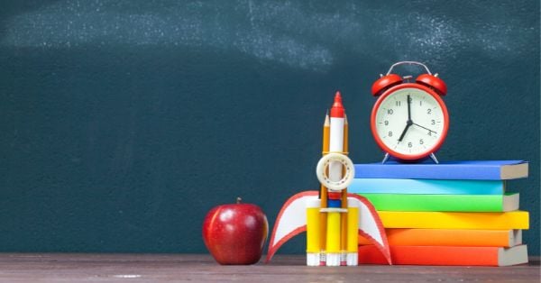 A red apple, a rocket model made of pencils, a stack of colorful books, and a red alarm clock sit on a desk in front of a green chalkboard—an inviting setup for independent learners in a Montessori-friendly classroom. Montessori Nature Printables