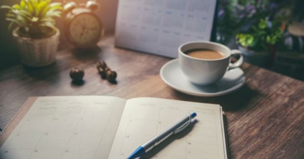 An open planner with a pen on a wooden table, next to a cup of coffee, a clock, potted plants, and a calendar—perfect for jotting down homeschooling tips or reflecting on homeschooling regrets. Montessori Nature Printables